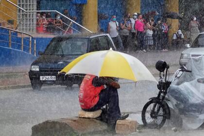 Un valet usa un paraguas para protegerse de un aguacero mientras la gente hace cola en la entrada de una tienda estatal de dólares en La Habana, Cuba, el viernes 11 de junio de 2021. (AP Foto/Ramon Espinosa)