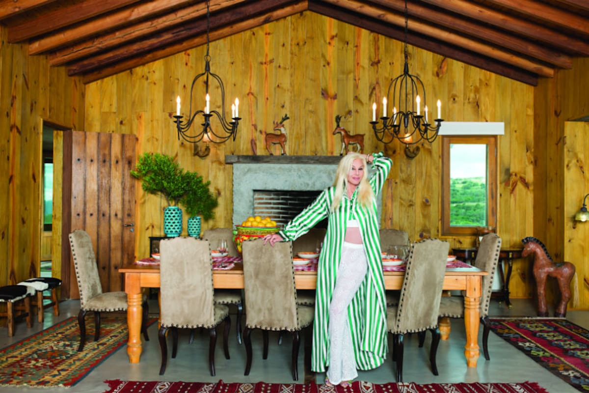 Un vista magnífica del salón comedor. Como en toda la casa, las paredes y el techo están revestidos en madera. La gran mayoría de los muebles de “La Tertulia” fueron importados desde los Estados Unidos. Susana posa con una camisola larga a rayas de Dolce & Gabbana, pantalón y