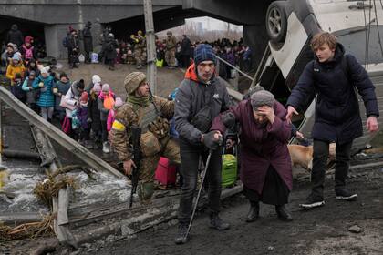 Una anciana recibe ayuda mientras cruza el río Irvin, bajo un puente destruido por un ataque aéreo ruso, mientras civiles huyen de la localidad de Irpin, Ucrania, el sábado 5 de marzo de 2022. (AP Foto/Vadim Ghirda)