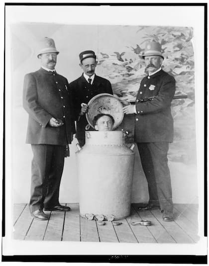 Una de las famosas demostraciones de escapismo de Harry Houdini. El mago aparece dentro de un gran contenedor metálico (un bidón de leche), con la cabeza apenas asomando por la tapa. A su alrededor, tres hombres —dos policías y un asistente— observan la escena. Este tipo de imagen se usaba como prueba pública de sus proezas, y muchas veces los policías participaban para verificar que no había trampa antes del encierro