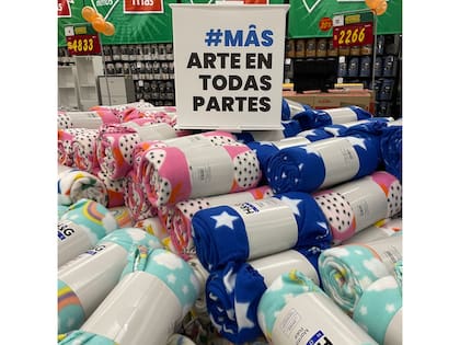 Una escena evoca las fotografías de la serie Pop Latino de Marcos López: un cubo de cartón anuncia el título del programa, “Mâs arte en todas partes” sobre una pila de mantas polares en venta