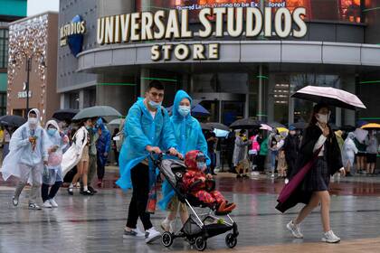 Una familia con impermeables y cubrebocas caminan frente a otros visitantes con cubrebocas formados para entrar a Universal Studios Beijing en Beijing, el 20 de septiembre de 2021. Miles de personas asistieron a la inauguración del parque temático a pesar de la lluvia. (Foto AP/Andy Wong)