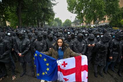 Una mujer sostiene una bandera de Georgia y una de la UE frente a agentes de la policía que impiden el paso a los manifestantes durante una protesta contra una ley cerca del Parlamento en el centro de Tiflis, Georgia, el martes 14 de mayo de 2024. (AP Foto/Zurab Tsertsvadze)