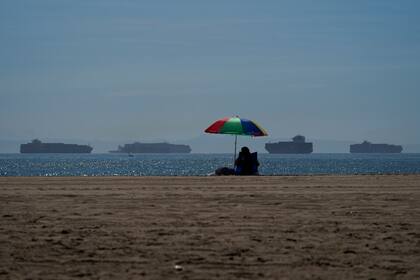 Una persona, sentada en la playa de Seal en California, el 1 de octubre de 2021, mientras mercantes esperan para atracar en los puertos de Los Ángeles y Long Beach en la distancia. (AP Foto/Jae C. Hong)