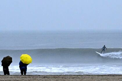 Unas personas caminan junto al mar mientras surfistas montan las olas durante una tormenta en Venice Beach, en Los Ángeles, California, el sábado 14 de enero de 2023. (Keith Birmingham/The Orange County Register via AP)