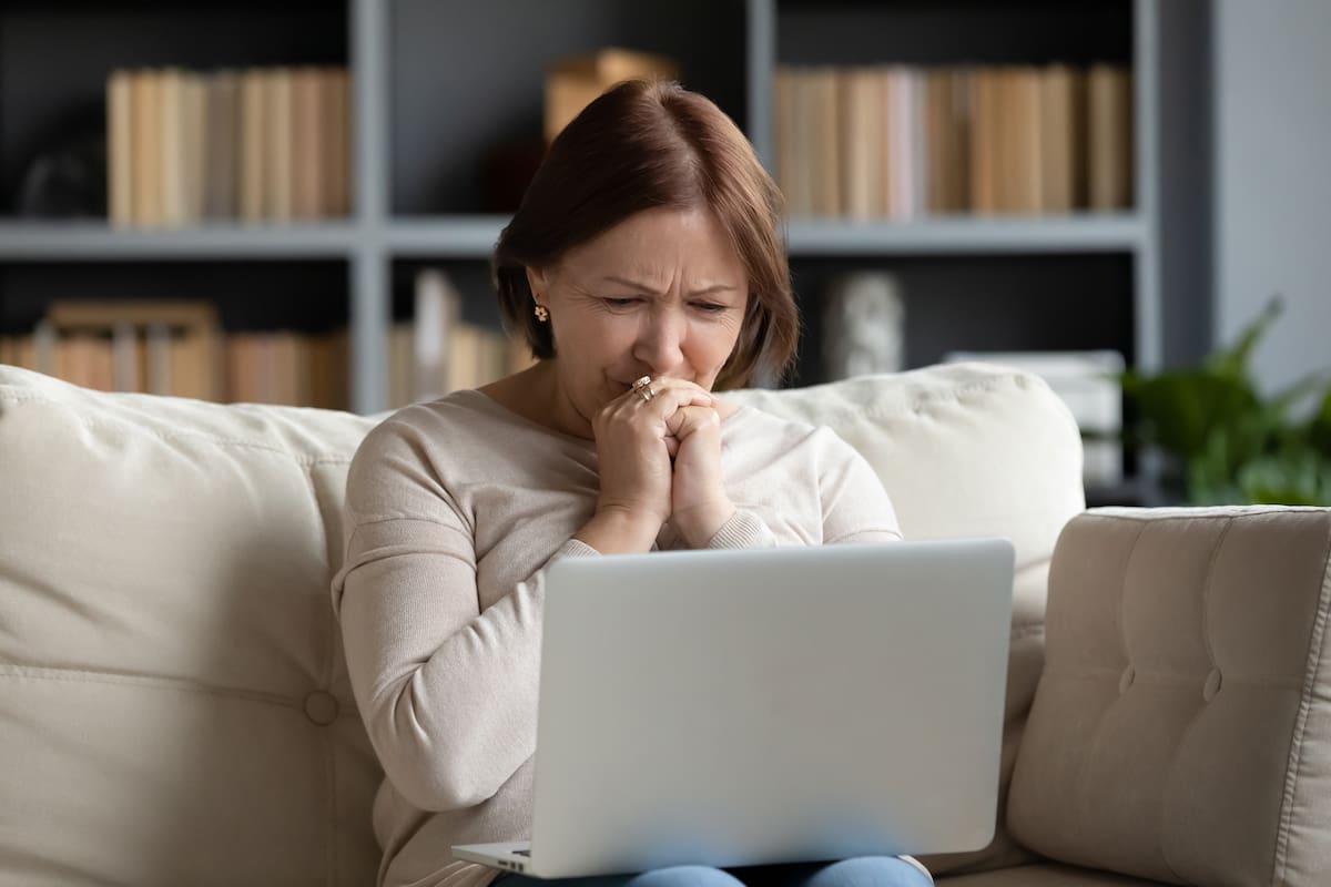 Upset senior woman sit on sofa in living room feel stressed crying reading bad unpleasant news on laptop, sad distressed middle-aged female worry get negative notice or email online on computer