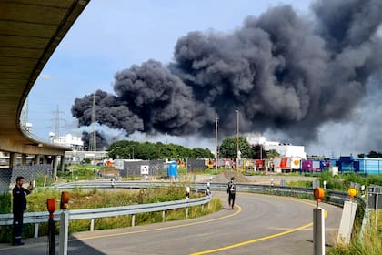 Vehículos de emergencias de bomberos, rescatistas y policías estacionados cerca del acceso al recinto industrial Chempark, sobre el que se alza una negra humareda, en Leverkusen, Alemania, el martes 27 de julio de 2021. (Oliver Berg/dpa via AP)