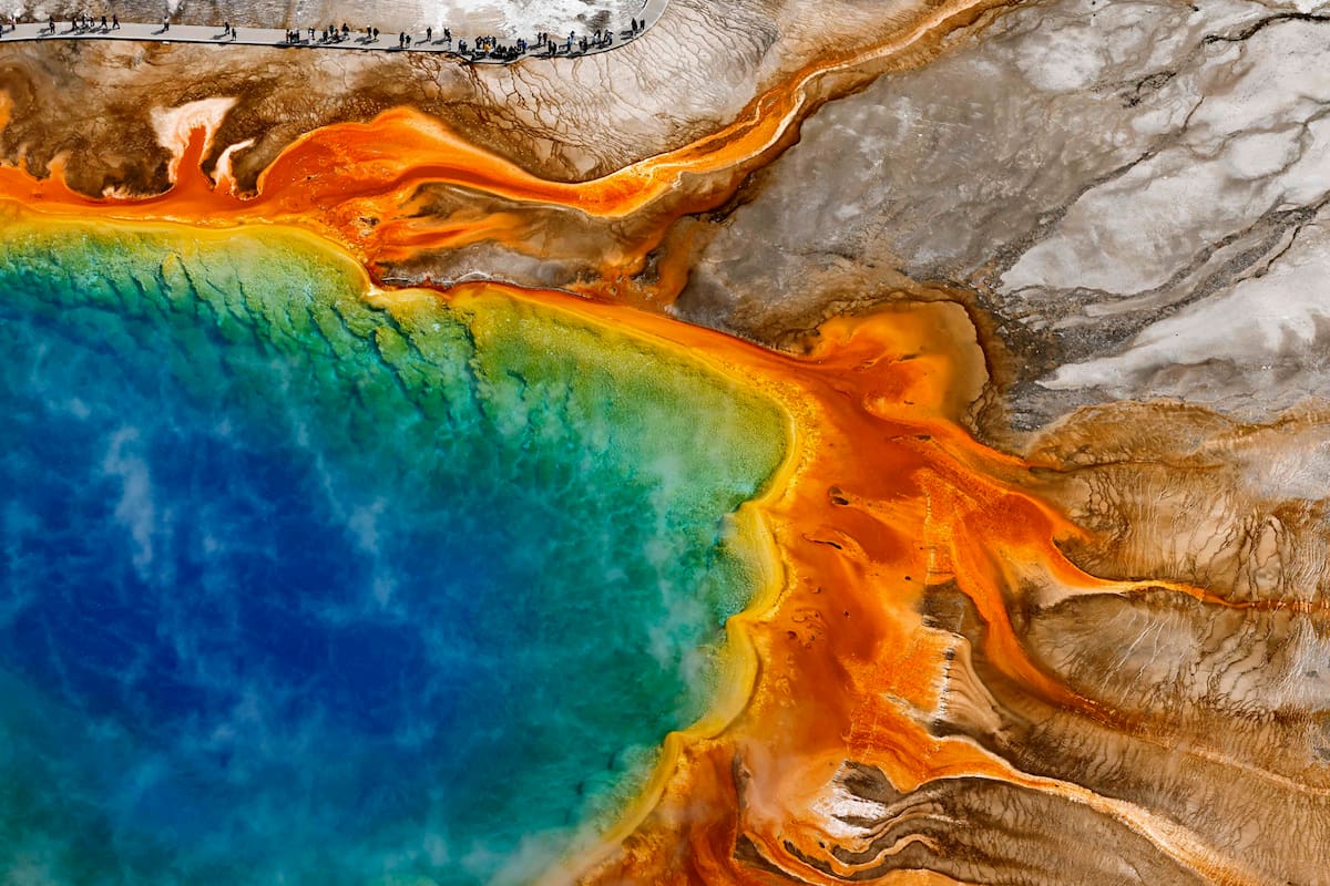 Vista aérea de visitantes caminando alrededor de Grand Prismatic Spring en la Cuenca de Géiseres de Midway el 24 de mayo de 2026 en el Parque Nacional de Yellowstone, Wyoming. Grand Prismatic Spring es la fuente termal más grande de los Estados Unidos, con más de 91 metros de ancho en su punto más amplio y más de 37 metros de profundidad. Los colores que se ven fuera del centro azul de la poza son creados por "termófilos", organismos microscópicos amantes del calor que proliferan en muchas de las fuentes termales de Yellowstone. El Parque Nacional de Yellowstone se convirtió en el primer parque nacional del país el 1 de marzo de 1872, cuando el presidente Ulysses S. Grant firmó una ley del Congreso que protegía la tierra "para el beneficio y disfrute del pueblo". Hoy, el parque de 2,2 millones de acres prospera como parte de uno de los últimos ecosistemas templados de gran tamaño casi intactos de la Tierra, famoso por conservar más de 10 000 formaciones hidrotermales y alrededor de 500 géiseres, aproximadamente el 50 % del total mundial. Con la mayor concentración de mamíferos en los 48 estados contiguos, Yellowstone es el único lugar en los EE. UU. donde los bisontes han vagado continuamente desde tiempos prehistóricos. Las comunidades nativas americanas habitaron y conservaron estas tierras durante más de 10 000 años, y 27 tribus individuales han sido reconocidas por el Servicio de Parques Nacionales por tener vínculos históricos y actuales con Yellowstone. La Semana de los Parques Nacionales celebrará el 250 aniversario de Estados Unidos con una celebración de una semana de duración en agosto que incluirá cientos de eventos conmemorativos en muchos de los 433 parques nacionales de Estados Unidos bajo el lema "Celebremos la historia de Estados Unidos". Mario Tama/Getty Images/AFP (Foto de MARIO TAMA / GETTY IMAGES NORTH AMERICA / Getty Images vía AFP)