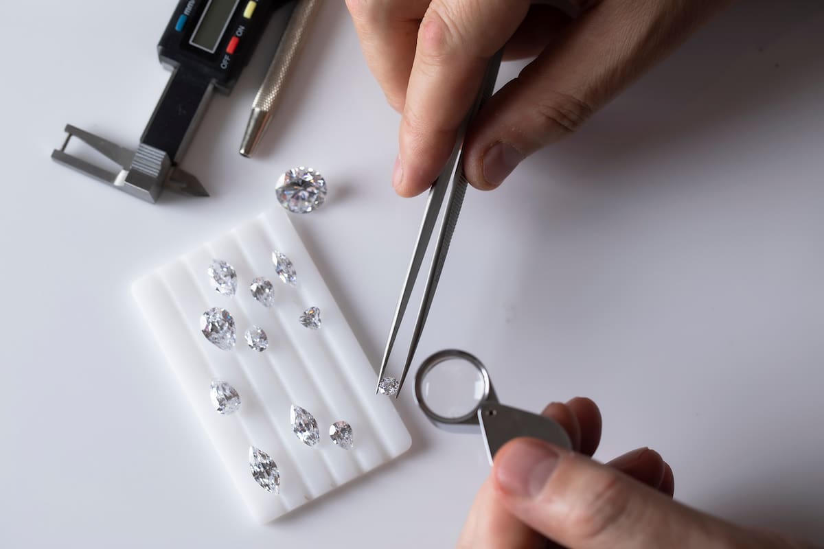 Workplace of jewellery buyer. Top view of hands with tools and diamonds of different cut on white platform for color determination.