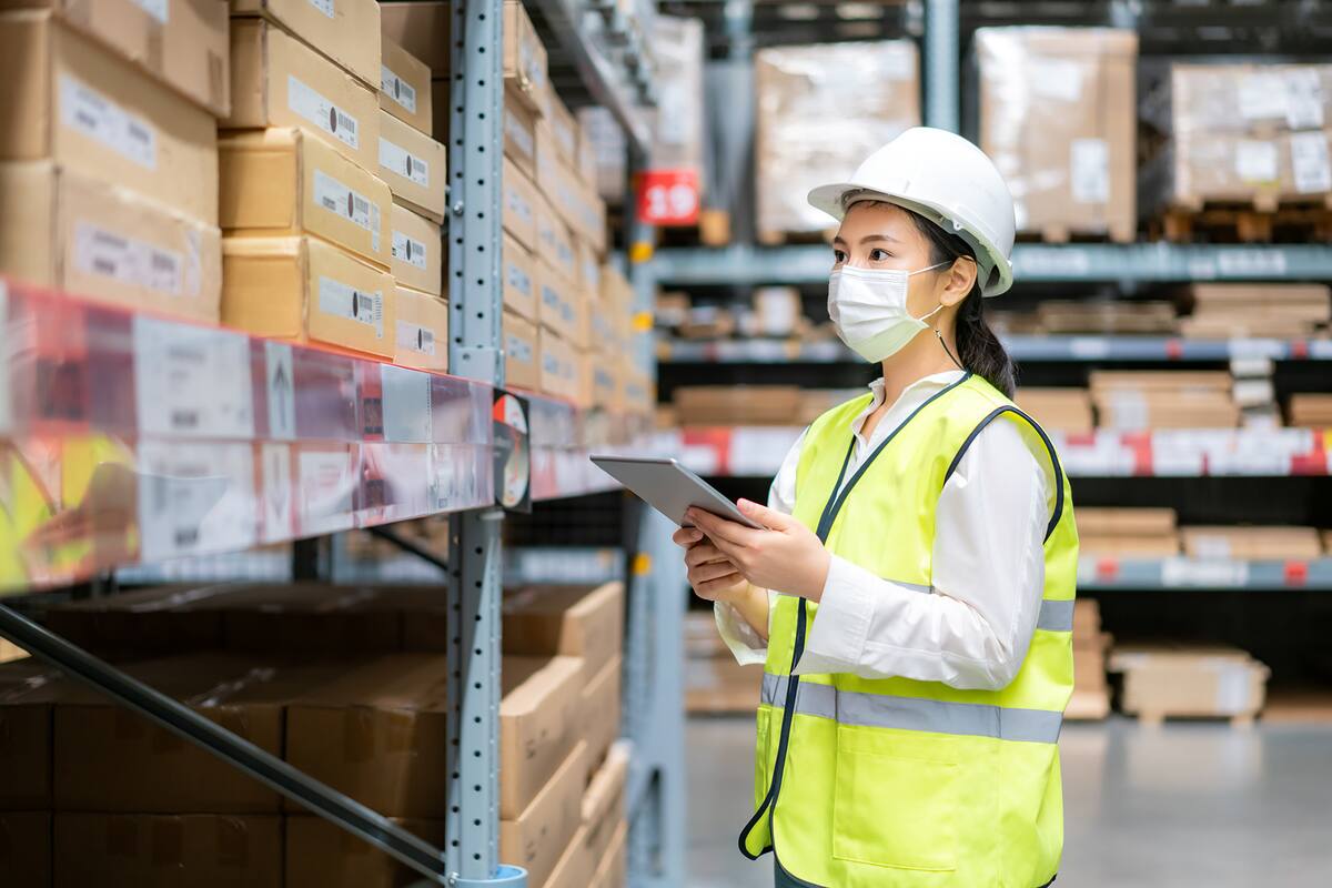 Young asian woman auditor or trainee staff wears mask working during the COVID pandemic in store warehouse shipping industrial. looking up and checks the number of items store by digital tablet.