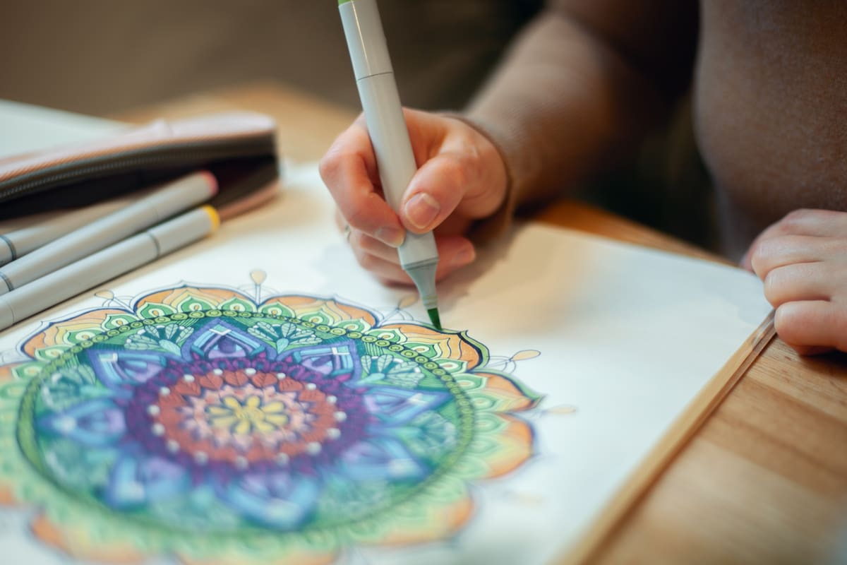 Young woman colouring mandala with markers and white rosary on table with cup of coffee near flower vase at home. Art therapy and meditation concept. Hobby and leisure during quarantine. Wellbeing.