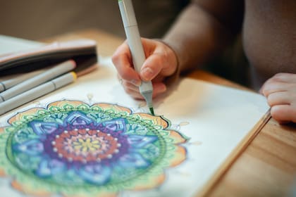 Young woman colouring mandala with markers and white rosary on table with cup of coffee near flower vase at home. Art therapy and meditation concept. Hobby and leisure during quarantine. Wellbeing.